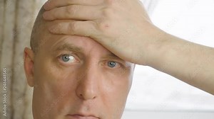 Adult white caucasian man rubs stroke bald head himself by hand at natural light backlit background. Portrait thinking man detail close up face. Male rubs his sore head. Extreme close up. Helth care