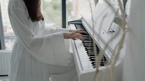 Woman in white robe playing piano in a bright room with large windows overlooking the city. Woman Playing Piano in Bright Room Wearing White Robe
