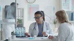 Young beautiful mixed raced female doctor showing x-ray image of brain on laptop screen and answering questions from patient during medical consultation in clinic