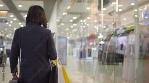Rear tracking shot of Asian woman walking through shopping mall with paper bags, then stopping by store display and taking photo of clothing on mannequin on smartphone