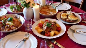 A table with a red tablecloth and a variety of food on plates. The table is set with forks, knives, and spoons, and there are wine glasses and cups. Scene is festive and inviting