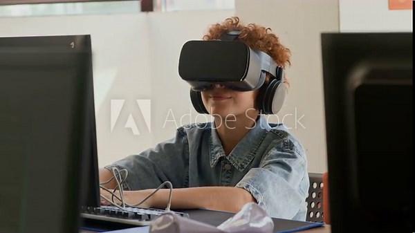 Cute schoolboy with virtual reality headset sitting by desk in front of computer monitor at lesson of programming, watching 3d video and commenting immersive experience