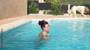 A woman is swimming in a pool with a dog watching her. The dog is brown and white
