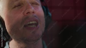 A close-up of a man's face passionately singing with his mouth wide open, wearing black studio headphones. The shot captures the intensity of the performance, set against a red background