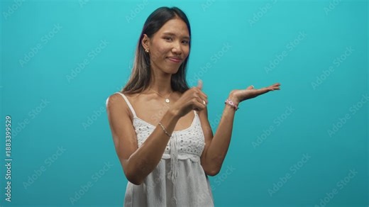 Young thai woman points to open palm while smiling and winking in a turquoise studio setting; playful demonstration.