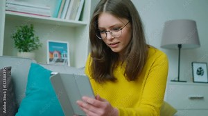 Teenager girl holding tablet computer while sitting on the sofa in the living room. Girl surfing the internet, watching videos and smiling. Close up