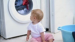 Little girl in white T-shirt sits on floor. Baby watches with interest how works washing machine drum. First year of life, knowledge of world around.