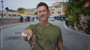Man wearing olive t shirt extends right hand for handshake toward camera on a cobbled street lined with blurred buildings and greenery; friendly greeting.