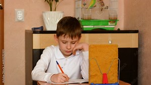 Caucasian boy of primary school age, sitting at the table, writes in a notebook, does his homework. The child learns to write in a notebook. The boy does school homework.