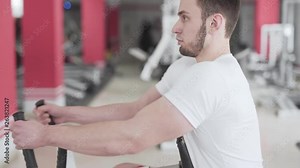 Close Up Footage of Young Caucasian Male in Sport Gym Using Rowing Machine with Both Hands and Looking Forward. Light Camera Movement and Blurred Gym Background