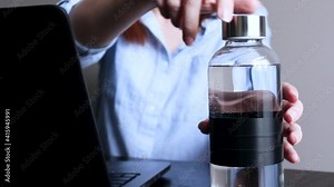 Woman works on a laptop, open water bottle, drinks water from a bottle. Focus on a glass bottle with clean water. Space for text.