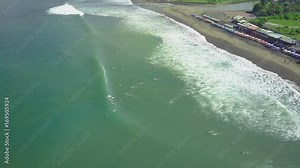 AERIAL: Beginner surfers learning to surf on surfboards and longboards at sunny surfspot. Crowds of surfers in the ocean catching breaking waves. People relaxing under sunshades on sandy beach in Bali