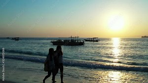 Two girls walking barefoot on wet sand of exotic beach enjoying beautiful sunset with low light reflecting on sea surface in Bali
