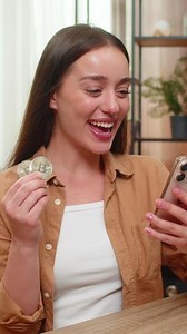 Young woman at home table watches smartphone price line after closing bitcoin sale and smiles with joy. Caucasian girl celebrates profit as mobile phone chart shows crypto growth and successful trade.