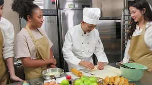 Cuisine course, senior male chef in cook uniform teaches young cooking class students to knead and roll pastry dough, prepare ingredients for bakery foods, fruit pies in stainless steel kitchen.