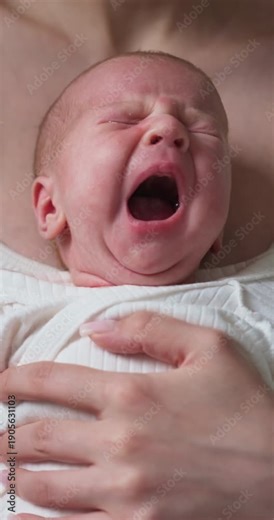 Close-up of a distressed newborn baby with eyes shut tight and mouth open wide in a loud cry, held by a parent's gentle hand