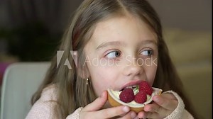 Adorable little girl eating fresh sweet raspberry cake indoors. Girl eating cake with cream and fruits. Child celebrates birthday with fresh raspberry tart