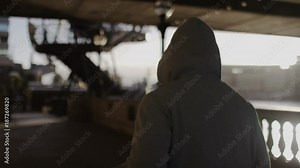 Camera follows a hooded jogger as they run by a river in the city, in slow motion