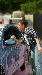 A beautiful young girl washes her car at the car wash using a sponge and cleaning solution.