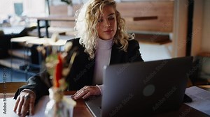 Attractive blonde hair female sits in front of open laptop computer, looks at screen and takes notes while working remotely. Modern young woman learns online with help of courses. Slow motion