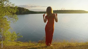 A beautiful girl in a long red dress stands near a forest lake and dances at sunset