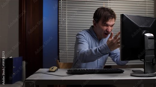 Frustrated man clenching fists and yelling at personal computer monitor in office, experiencing anger and stress due to technical issues or work overload