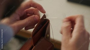 Leather craftsman works in his workshop. Master making leather wallet. Man uses plywood stitching pony or wooden vise for leatherworking. Guy with a needle and thread sewing goods in atelier.