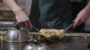 This slow motion video shows the hands of a chef flipping a okonomiyaki pancake on a grill.