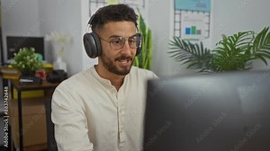 Young hispanic man having a videoconference indoors in an office setting, wearing headphones and glasses, surrounded by plants and charts, engaged in work activities.