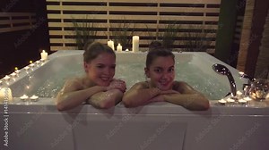 Two happy girls who came to get a spa treatment, enjoy a jacuzzi. Girls spend time in the water with aromatic oils, which moisturize the skin. Romantic atmosphere, candles, nice music help to relax.