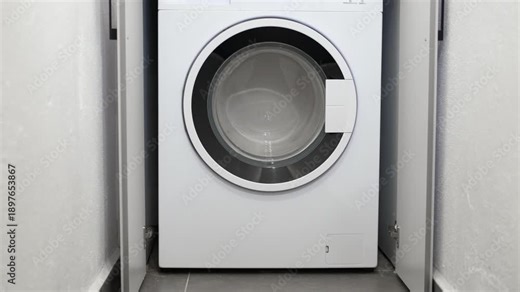 Person closing the cabinet door on a modern washing machine. Modern white washing machine built into a minimalist cabinet in a laundry room, with a person approaching and closing the cabinet doors