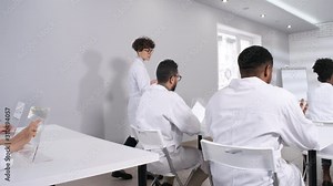 Medium shot of female university teacher in white lab coat walking through classroom while giving lecture, checking on students working on futuristic touchscreen devices. Template for AR, VR effects.