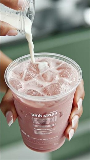 A close up of a brown woman’s hand pouring a iced pink latte in a transparent plastic cup with te...