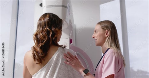 Female tech guiding female patient into mammogram position at clinic, adjusting gown aligning torso