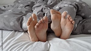 Closeup as the feet of a man and a woman lie entwined under the blanket on a soft bed. Feelings of warmth, love, and affection in a cozy home setting