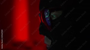 Hacker in a mask and glasses hacks into the system, a code with a running line is reflected in the glasses close-up, a red disturbing background, cyberattack