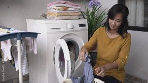 happy asian woman in front of the washing machine doing some laundry loading clothes inside