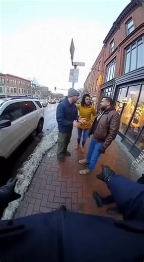 Police body-cam footage captures officers actively de-escalating a heated argument outside a storefront. Voices are raised, one person gesturing aggressively while an officer stands between them with palms out, issuing calm but firm commands. The body cam audio picks up tense breathing and overlapping voices as the officer works to keep distance and control the scene. Suddenly, the sound of screeching tires cuts through the confrontation. A car slams into another vehicle in the street, forcing i