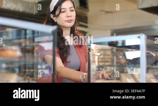 Woman 30s wearing headband, rust shirt, reaching through glass display, serving pastry to customer. Bakery, cafe, staff, counter, reflections, bokeh, warmth Stock Video Footage - Alamy
