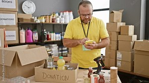 Bearded, middle-aged, handsome caucasian man is a dedicated volunteer at the charity center, engrossed in checking products using his smartphone, lending a helping hand in the community.