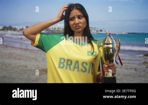 Woman wearing brazil t shirt holds trophy and scratches head on a sunny beach showing puzzled facial expression; confusion defeat Stock Video Footage - Alamy