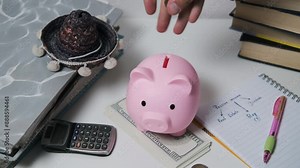 Hand drops coins into a pink piggy bank that sits on top of a stack of dollar bills. A calculator, a notepad, and a pen are also present. Savings, financial security and budgeting concept