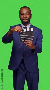 Vertical video: Front view of businessman filmmaker holding filming slate on camera, using flapperboard in movie making and post production industry. Confident person in formal suit posing over green