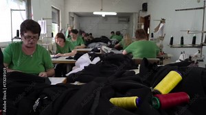 Colorful spool threads on the black fabric textiles in focus, group of female tailors stitching at sewing machines in clothing factory in the blurred background, dolly shot, camera motion to the women
