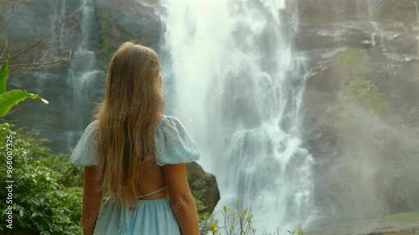Woman with long hair standing in front of majestic waterfall surrounded by lush greenery, appreciating beauty of nature, wearing light blue dress, back view. Travel and Adventure.