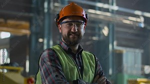 Professional heavy industry employee in protective uniform, helmet and goggles stands at the manufacturing factory. Male technician or engineer smiles and looks at camera. Assembly plant. Portrait.
