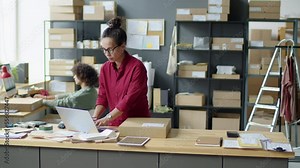 Woman using laptop and writing address on box label while preparing parcel for shipping in delivery service office