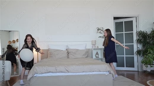 Two children, happy laughing two cute girl having fun at pillow fight with feathers in the air jumping, laughing and giggling in a white bedroom with pastel light bedding