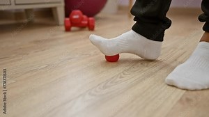 A man performs foot exercises using a red ball on a wooden floor at a physiotherapy clinic.