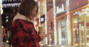 Woman using her Mobile Phone in the street, night light Background. Girl texting on smart phone and drink coffe.